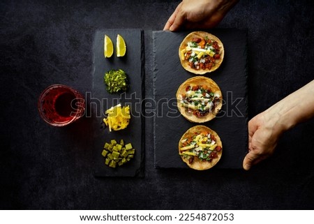 Mexican tacos served on black table near various toppings, top view of crop anonymous person serving tacos on black slate board with assortment of toppings and drink on table