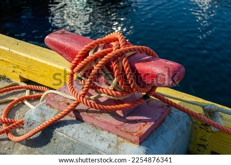 A rusty red metal mooring or bollard for boats on a yellow wooden wharf with a red colored braided fishing rope. The rope is tangled around the mooring. There's water in the background.