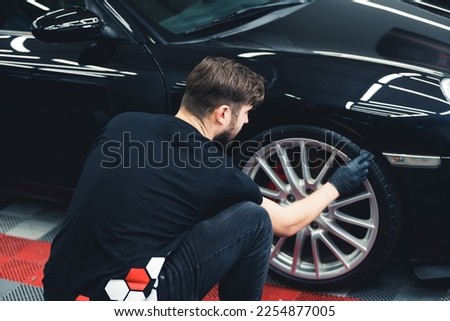 Rear view of caucasian man wearing black gloves crouched blackening tires of car using sponge. Professional car detailing in a garage. Horizontal indoor shot . High quality photo