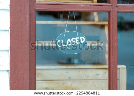 A small green round shaped wooden business closed sign with beige painted letters hangs in a glass window of a red wooden building. The word closed is handwritten on the circular handpainted sign.  