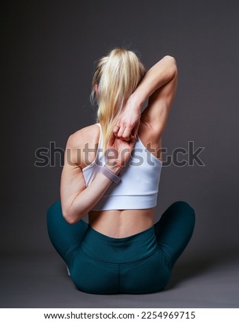 A European, young girl with blond hair in a white T-shirt and green pants is doing yoga. Hands in the lock behind the back. On a gray background. Athletic body.