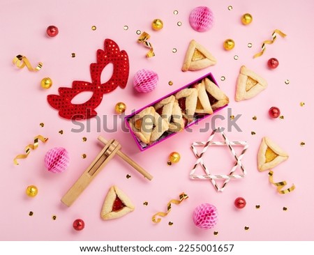 Purim celebration jewish carnival holiday concept. Tasty hamantaschen cookies, Triangular pastry, red carnival mask, noisemaker, sweet candies and party decor on pink background, Top view, copy space