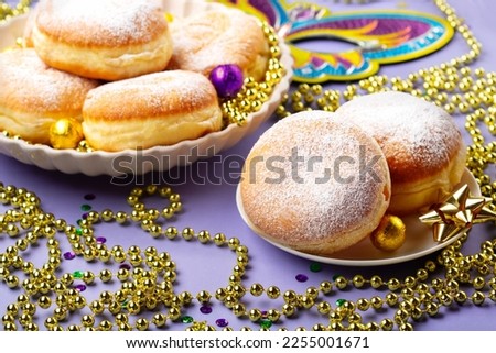 Mardi Gras King Cake doughnuts or donuts, masquerade festival carnival mask, gold beads and golden, green confetti on purple background. Holiday party invitation, greeting card concept. Royalty-Free Stock Photo #2255001671