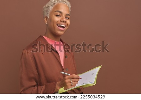 Young cheerful attractive African American woman zoomer with notebook and pen in hands looking at camera with inspiration preparing to draw creative sketch of future picture stands on brown background