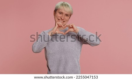 Woman in love. Smiling woman with short blonde hair makes heart gesture demonstrates love sign expresses good feelings and sympathy. Young girl isolated on pink background. Lgbt gay lesbian people