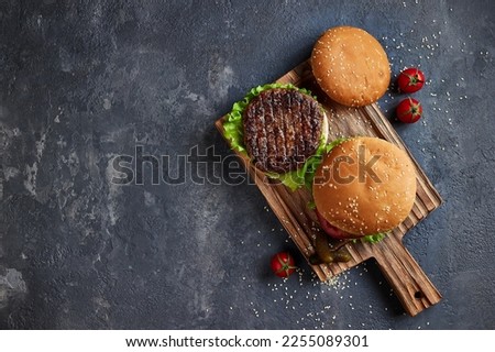 Tasty homemade grilled burger with beef, cheese, tomatoes, pickles, onion and lettuce on a dark concrete background with copy space. Top view flat lay. Fast food and junk food.