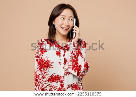 Young woman of Asian ethnicity wear red shirt talk speak on mobile cell phone conducting pleasant conversation isolated on plain pastel light beige background studio portrait. People lifestyle concept