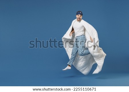 Full length overjoyed funny young man 20s wearing pajamas jam sleep mask resting relaxing at home jump high wrap with duvet blanket isolated on dark blue background. Good mood night bedtime concept. Royalty-Free Stock Photo #2255110671