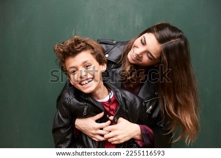 beautiful female with long hair and cute boy wearing black leather jackets hugging and laughing in studio, emerald green wall on background. Siblings having fun together