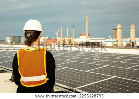 Female solar installer inspecting a commercial rooftop solar system in Charlestown, Boston, Massachusetts, U.S. with a wind turbine and a natural gas electric generation facility in the background.