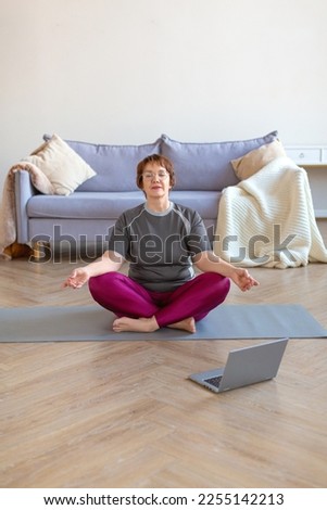 An elderly woman meditates at home in a lotus position in front of a laptop monitor. The concept of a healthy and active lifestyle in old age. Vertical photo.
