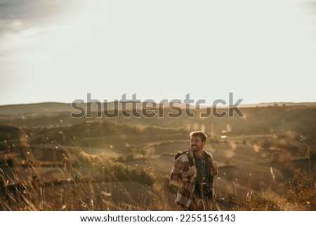 Happy man walking and carrying his backpack and hiking. He is smiling and touching the grass. He is going through a grass meadow.