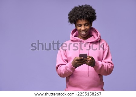 Young happy cool curly African American teen guy wearing pink hoodie holding cell phone using mobile digital apps on cellphone technology texting on smartphone isolated on light purple background.