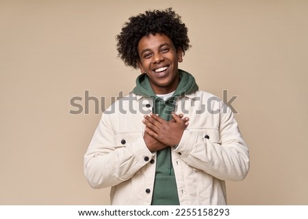Happy pleased excited grateful African American teen guy holding hands on chest expressing gratitude, feeling trust hope love in heart, impressed with kindness isolated on beige background. Royalty-Free Stock Photo #2255158293