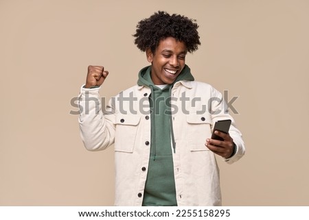 Happy African American teenager, ethnic cool guy student winner holding cell phone winning money online on cellphone, using mobile app celebrating great result with yes isolated on beige background.