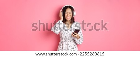 Beautiful asian woman listening music in headphones, using smartphone streaming app and smiling, standing over pink background.