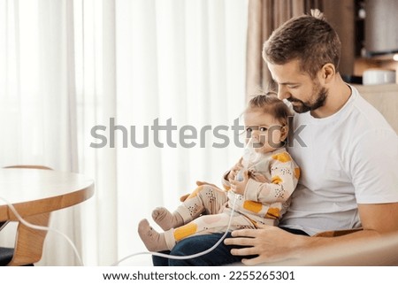 A daughter is using nebulizer by her own while her father is holding her in his lap. Royalty-Free Stock Photo #2255265301