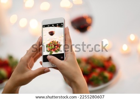 Cropped of lady taking photo of glass with red wine and plate full of strawberry on festive table decorated with lit candles, woman sharing photos on social media while romantic dinner, using phone