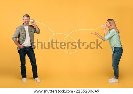 Communication and fun. Happy middle aged spouses talking through tin can telephone, standing over yellow studio background. Man and woman hearing and speaking through cups Royalty-Free Stock Photo #2255280649