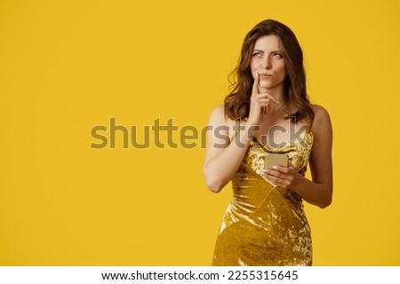 Pensive adorable caucasian brunette woman looking aside, touching chin and holding smartphone, thinking about present, posing over yellow studio background, copy space