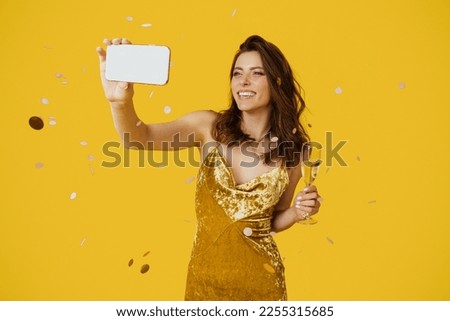 Happy woman in stylish dress taking selfie with glass of champagne, standing over yellow studio background with falling confetti. Positive lady making photo of herself during fun party