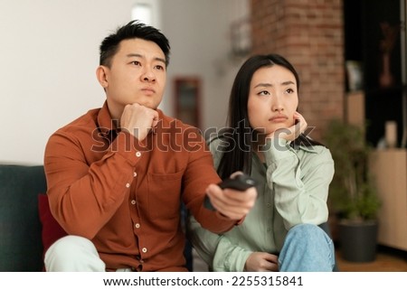 Discontented asian couple watching television, pointing remote controller and switching TV channels, sitting on sofa at home. Boring movie, displeased viewers concept