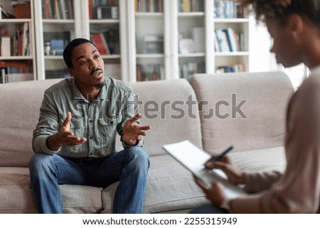 Emotional african american millennial guy in casual sitting on couch at therapist office, sharing feelings and gesturing, young black man attend therapy session with woman psychologist, free space Royalty-Free Stock Photo #2255315949