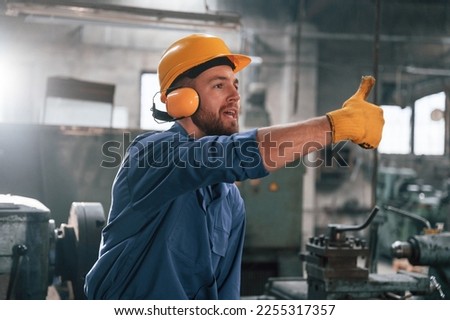 Man in hard hat and headphones is showing thumb up. Factory worker in blue uniform is indoors.