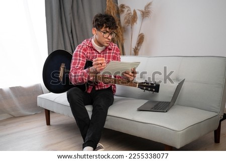 Young arabic composer man composing song using guitar and laptop at home sitting on sofa. Copy space