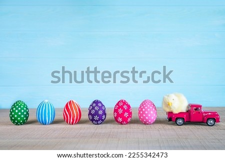Easter composition with yellow fluffy fledgling chicken at pink car and colored easter eggs against the blue wooden background. Easter card Royalty-Free Stock Photo #2255342473