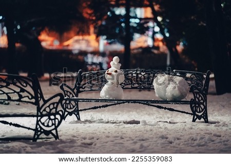 Lonely snowman on a bench. A beautiful snow man created by a child is standing on a bench in a park during winter evening.