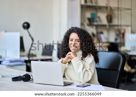 Young happy latin business woman company worker sitting at desk working on laptop. Smiling female professional executive hr manager using computer in corporate office looking at camera. Portrait. Royalty-Free Stock Photo #2255366049