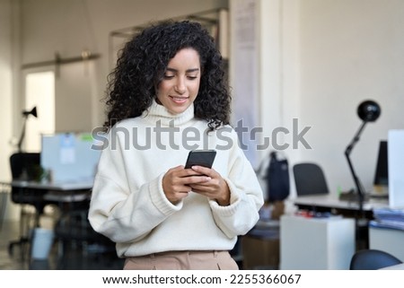 Smiling young professional business woman, happy corporate leader holding cellular smartphone working standing in office using mobile applications cell phone technology device looking at cellphone. Royalty-Free Stock Photo #2255366067