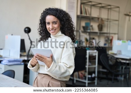 Smiling latin young professional business woman corporate marketing manager, female worker holding digital tablet computer fintech tab at work standing in modern company office looking at camera. Royalty-Free Stock Photo #2255366085