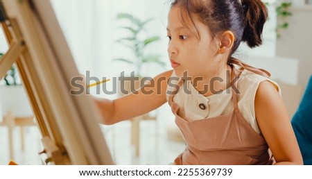 Close-up Asian toddler girl with apron sit on sofa chair in front of painting canvas with color palette use paint brush focus on painting abstract on weekend at home. Creative lifestyle kid concept.