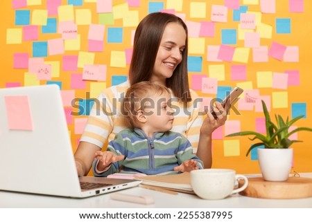 Photo of smiling woman with brown hair wearing striped t-shirt sitting at table against yellow wall with colorful sticky notes, mother holding cell phone and playing with baby while being on office.
