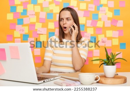 Photo of shocked dark haired woman wearing striped t-shirt sitting at table, posing against memo cards on yellow wall, working on laptop in office, talking cell phone, keeps mouth open.