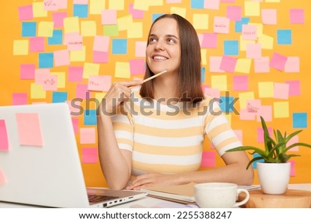 Image of pensive dreaming smiling woman with brown hair wearing t-shirt sitting at table posing against yellow wall with adhesive small notes, sits in front of laptop and thinking about something.