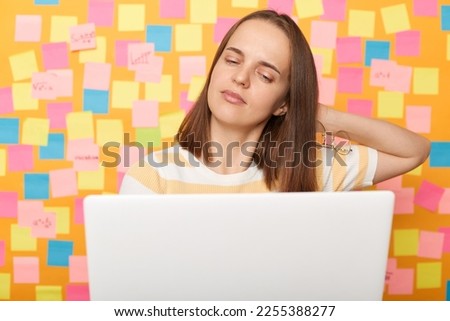 Portrait of tired sad sleepy woman wearing striped T-shirt posing over sticky notes to write reminder on yellow wall, working on pc computer, being overworked, feels pain in neck.