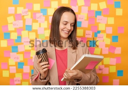 Image of smiling beautiful woman wearing beige jacket posing over sticky notes to write reminder on yellow wall, reading something in organizer, drinking hot tasty takeaway coffee.