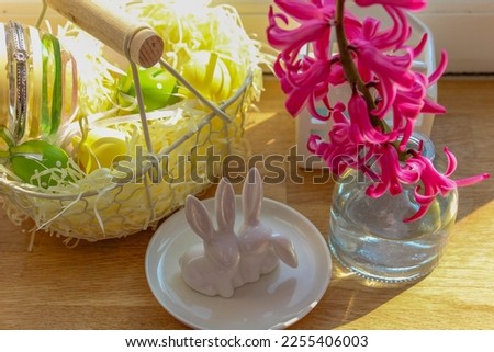 A rustic basket, white ceramic Easter bunnies and a hyacinth in a glass vase on a wooden windowsill bathed in sunlight. Easter decor. High quality photo