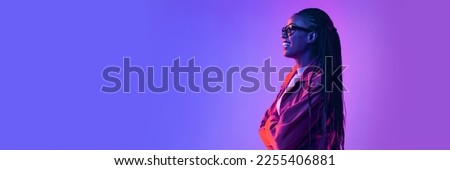Cheerful african woman in glasses posing, looking away with smile over gradient blue purple background in neon light. Banner, flyer. Concept of emotions, facial expression, sales, ad, fashion