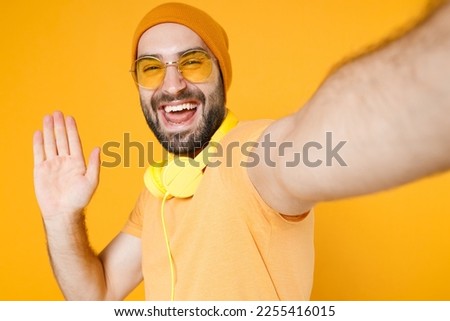 Close up of laughing young bearded man wearing basic casual t-shirt headphones eyeglasses hat doing selfie shot on mobile phone waving greeting with hand isolated on yellow background studio portrait