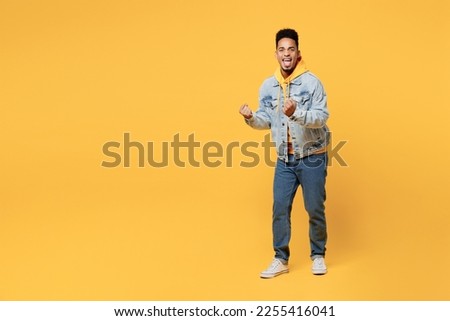 Full body side view young man of African American ethnicity wear denim jacket hoody look camera do winner gesture celebrate clenching fists say yes isolated on plain yellow background studio portrait
