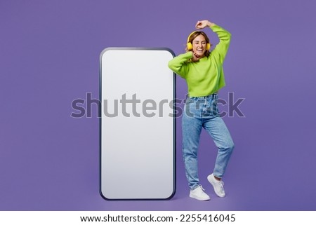 Full body young woman she wear casual green knitted sweater headphones stand near big huge blank screen mobile cell phone with area listen to music isolated on plain pastel purple background studio