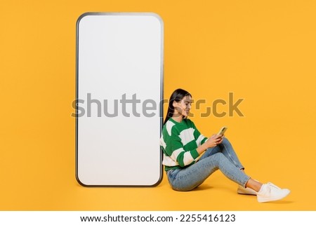 Full body young latin woman 30s wear casual cozy green knitted sweater big huge blank screen mobile cell phone smartphone with area use smartphone isolated on plain yellow background studio portrait