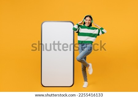 Full body young latin woman wear casual green knitted sweater headphones big huge blank screen mobile cell phone smartphone with area listen music isolated on plain yellow background studio portrait