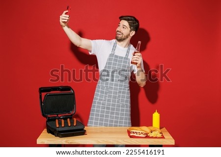Young housekeeper chef cook baker man in grey apron work at table with grill kitchenware doing selfie shot on mobile cell phone hold beer isolated on plain red background Process cooking food concept
