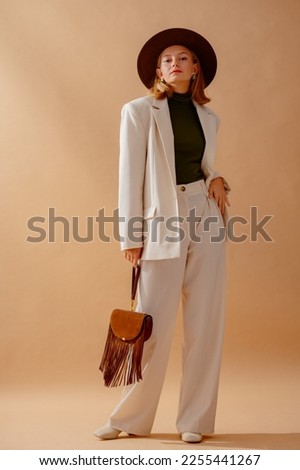 Fashionable confident woman wearing elegant white suit with blazer, wide leg trousers, turtleneck top, hat, leather ankle boots, holding brown suede fringed bag,  posing on beige background Royalty-Free Stock Photo #2255441267