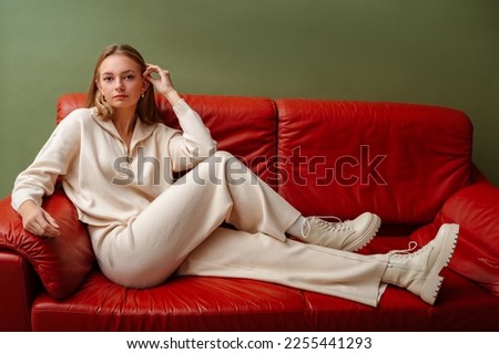 Fashionable confident woman wearing elegant white knit suit, with zip neck sweater, wide leg trousers, leather ankle lace up boots, sitting, posing on leather couch. Copy, empty space for text Royalty-Free Stock Photo #2255441293